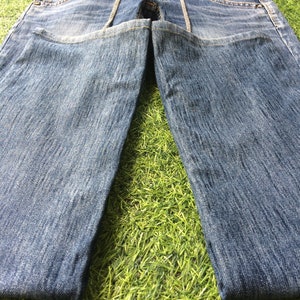 May include: Faded light blue denim jeans. The jeans feature a classic five-pocket design, a zip fly, and decorative stitching along the front pockets and seams. The jeans are displayed on a green surface.