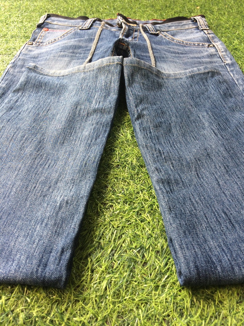 May include: Faded light blue denim jeans. The jeans feature a classic five-pocket design, a zip fly, and decorative stitching along the front pockets and seams. The jeans are displayed on a green surface.