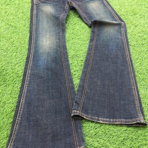 May include: Dark wash denim jeans with a flared leg design. The jeans feature classic five-pocket styling, a button and zipper closure, and contrasting tan stitching. The denim has a faded, worn look, and the jeans are laid out on a green surface.