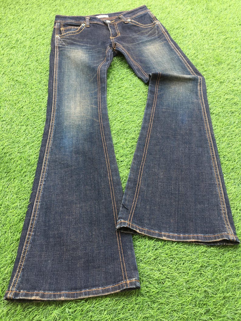 May include: Dark wash denim jeans with a flared leg design. The jeans feature classic five-pocket styling, a button and zipper closure, and contrasting tan stitching. The denim has a faded, worn look, and the jeans are laid out on a green surface.