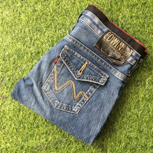 May include: Blue denim jeans with distinctive pocket detailing and gold stitching. The jeans have a black leather patch with "EDWIN" and "503" printed on it. The jeans are folded on a green grassy surface.