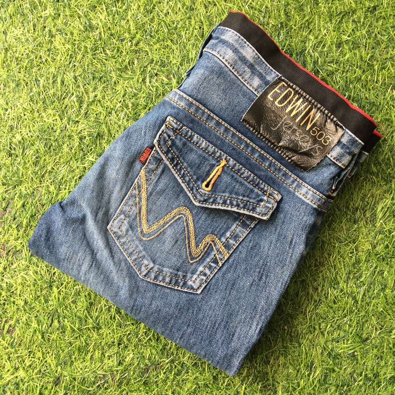 May include: Blue denim jeans with distinctive pocket detailing and gold stitching. The jeans have a black leather patch with "EDWIN" and "503" printed on it. The jeans are folded on a green grassy surface.
