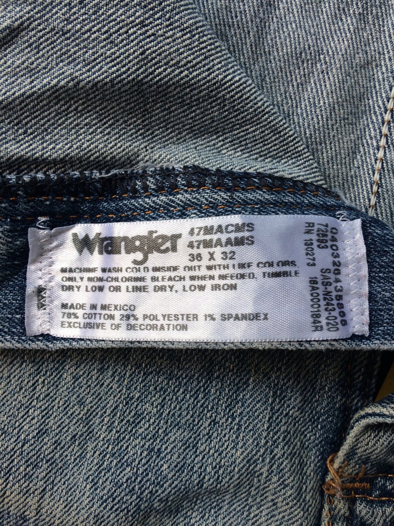 May include: A white Wrangler jeans care label with blue text. The label states "47MACMS 47MAAMS 36 X 32" and includes washing instructions. The label also states "Made in Mexico 78% Cotton 29% Polyester 1% Spandex Exclusive of Decoration."