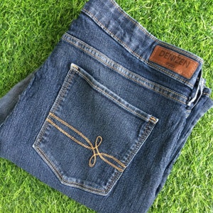 Può includere: Jeans in denim blu scuro con una tasca posteriore con una toppa in pelle marrone con il testo "DENIZEN" e un design cucito.