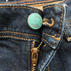 May include: Close-up of a pair of dark blue denim jeans. The jeans feature a brass zipper and a turquoise button with a decorative pattern. The stitching is a contrasting brown color.