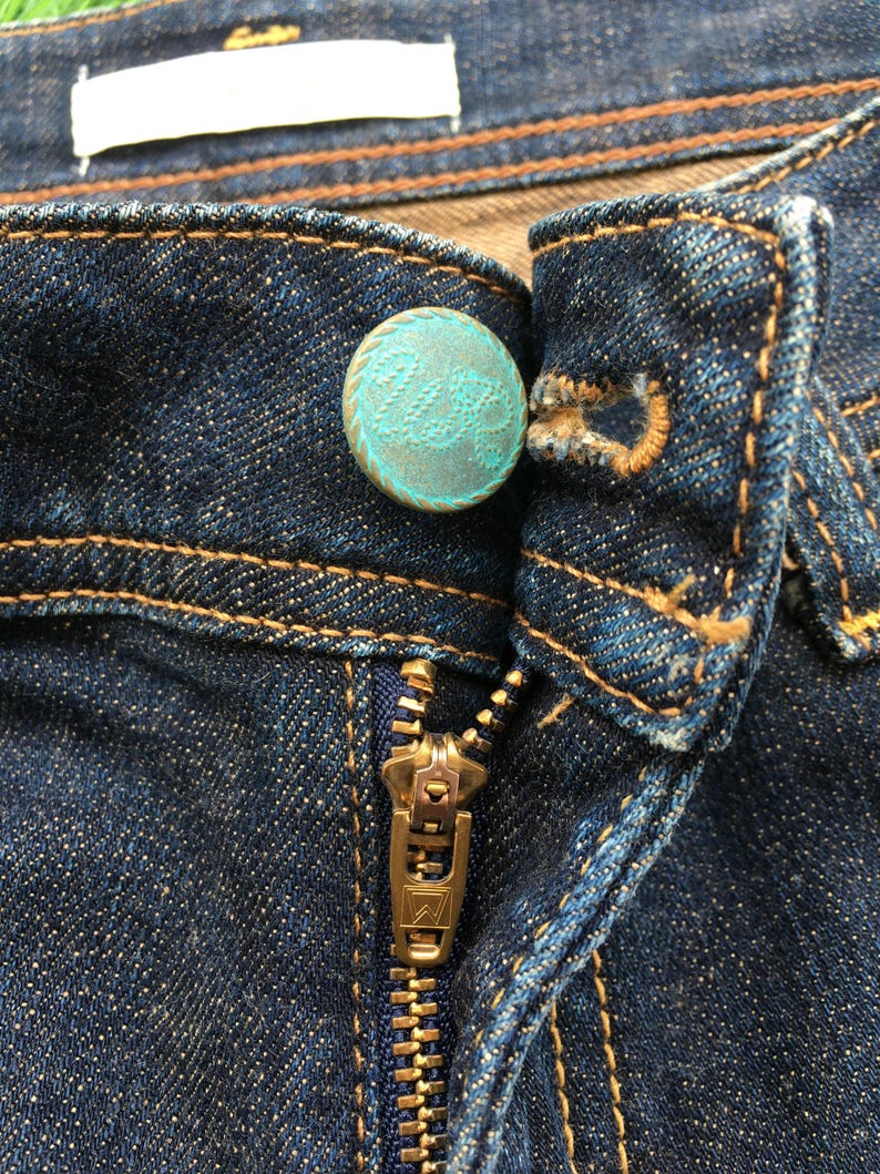 May include: Close-up of a pair of dark blue denim jeans. The jeans feature a brass zipper and a turquoise button with a decorative pattern. The stitching is a contrasting brown color.