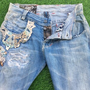 Puede incluir: Vaqueros de mezclilla azul desgastados con un adorno decorativo en el bolsillo delantero. Los vaqueros tienen un lavado azul claro y están rotos y desteñidos.