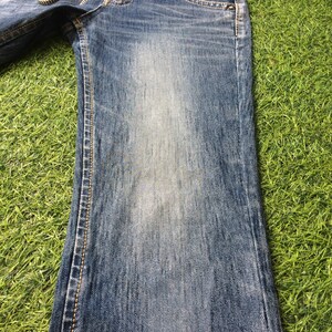 May include: Faded blue denim jeans with a slight flare at the hem. The jeans have a chain detail at the waist and contrasting stitching along the seams. The denim has a worn appearance.
