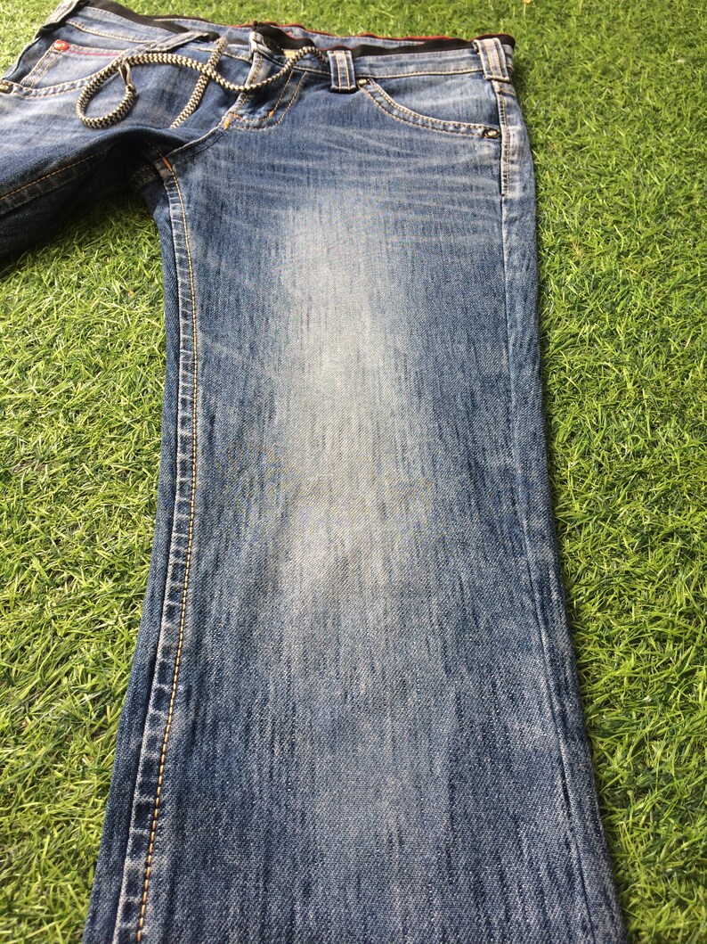 May include: Faded blue denim jeans with a slight flare at the hem. The jeans have a chain detail at the waist and contrasting stitching along the seams. The denim has a worn appearance.
