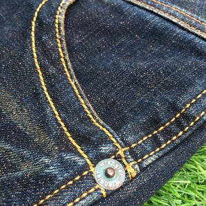 May include: Close-up of a section of dark blue denim jeans. The jeans feature yellow stitching, a turquoise button, and a turquoise rivet. The Wrangler logo is visible on the button. The fabric has a textured appearance.