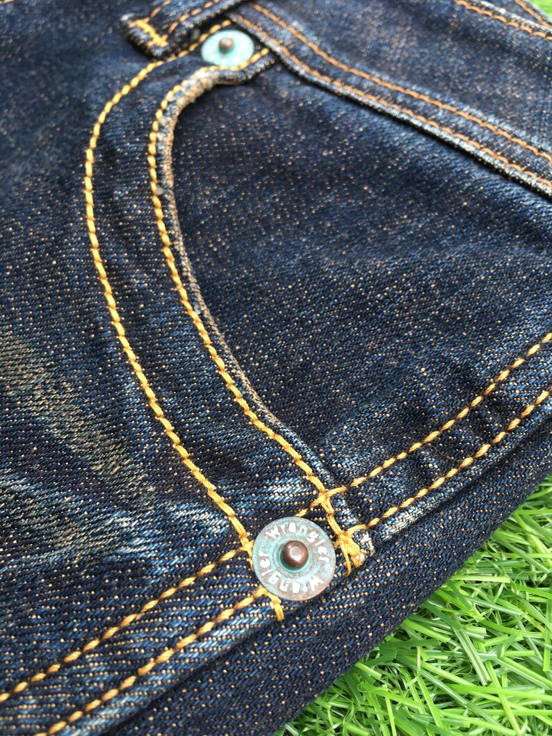 May include: Close-up of a section of dark blue denim jeans. The jeans feature yellow stitching, a turquoise button, and a turquoise rivet. The Wrangler logo is visible on the button. The fabric has a textured appearance.