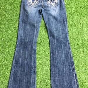 Op de afbeelding: Een paar blauwe denim jeans met een bootcut-been. De jeans heeft witte borduursels op de achterzakken en gouden knopen.