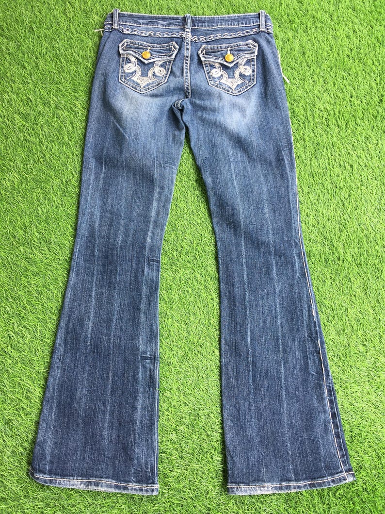 Op de afbeelding: Een paar blauwe denim jeans met een bootcut-been. De jeans heeft witte borduursels op de achterzakken en gouden knopen.
