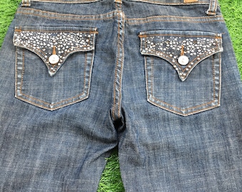 May include: Dark wash denim jeans with embellished back pockets. The pockets feature a decorative pattern of small, white, sparkling embellishments. The jeans have a classic five-pocket design with a button and zip closure.