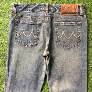 May include: A pair of blue denim jeans with a brown leather patch that says "Cecil McBee Denim Jeans". The jeans have two back pockets with a unique stitching design.