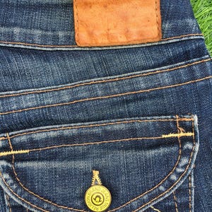 May include: Close-up of dark blue denim jeans with a brown leather patch and a pocket with a gold button. The jeans have orange stitching and a decorative design on the pocket.