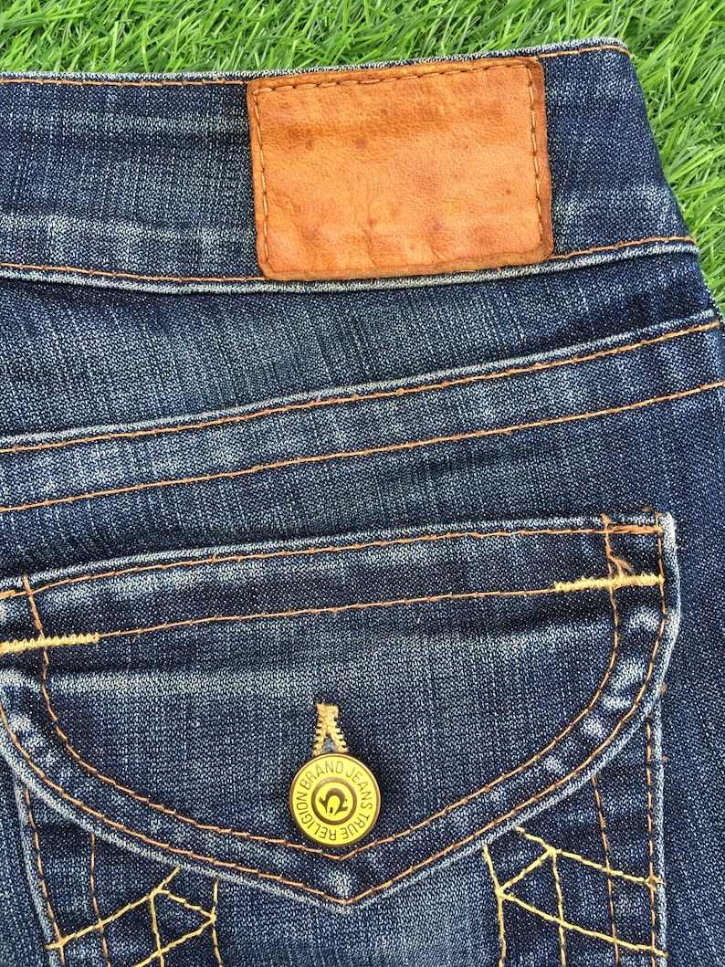 May include: Close-up of dark blue denim jeans with a brown leather patch and a pocket with a gold button. The jeans have orange stitching and a decorative design on the pocket.