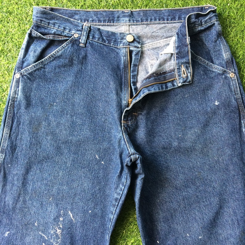 May include: A pair of blue denim jeans with a zipper fly and a button closure. The jeans have a worn look and have white paint splatters on them.