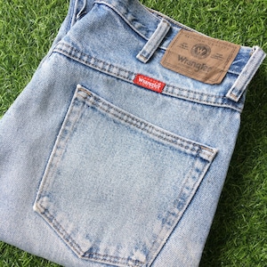 May include: Light blue denim jeans with a back pocket and a brown leather Wrangler label. The label has the Wrangler logo and the text "Premium Quality" and "Authentic Denim Wear".