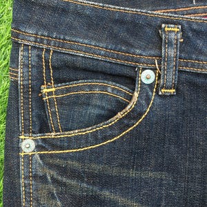 May include: Close-up of dark wash denim jeans. The jeans feature a small pocket with a curved opening, a button, and a belt loop. The stitching is a contrasting yellow-orange color.