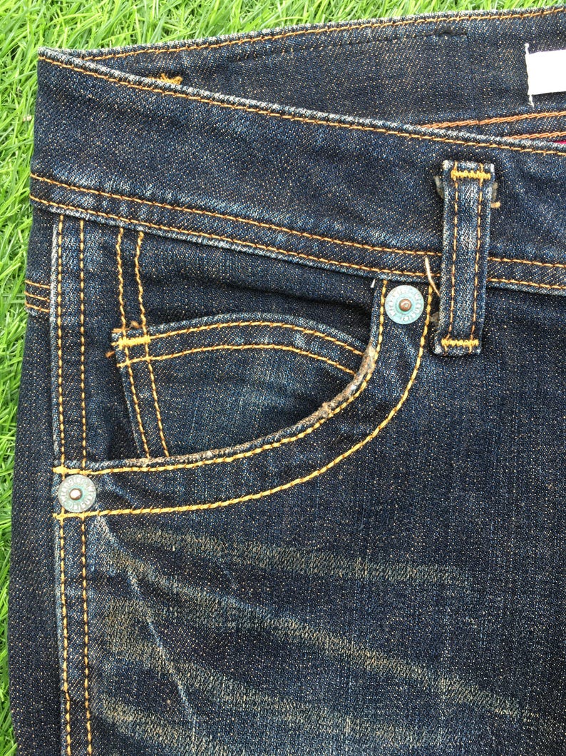 May include: Close-up of dark wash denim jeans. The jeans feature a small pocket with a curved opening, a button, and a belt loop. The stitching is a contrasting yellow-orange color.
