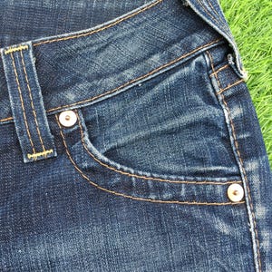 May include: Close-up of a pair of blue denim jeans. The jeans feature a classic five-pocket design with a small pocket on the front and two pockets on the back. The jeans have a button and zipper closure and are stitched with orange thread.