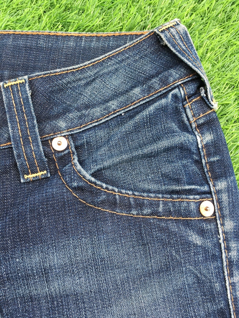 May include: Close-up of a pair of blue denim jeans. The jeans feature a classic five-pocket design with a small pocket on the front and two pockets on the back. The jeans have a button and zipper closure and are stitched with orange thread.