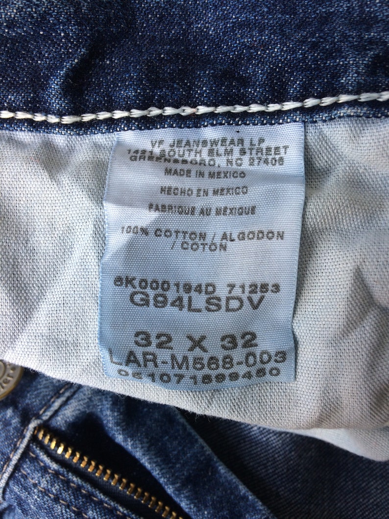 May include: A blue denim jean label with white stitching. The label has white text on a blue background with the text "VF Jeanswear LP 1481 South Elm Street Greensboro, NC 27406 Made in Mexico Hecho en Mexico Fabrique au Mexique 100% Cotton/Algodon/Coton 6K000194D 71253 G94LSDV 32 X 32 LAR-M568-003".