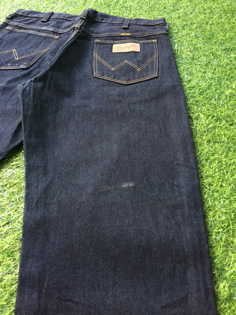 Peut inclure: Un jean en denim bleu fonc&eacute; avec un &eacute;cusson en cuir brun sur la poche arri&egrave;re qui dit "Wrangler".