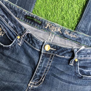 Op de afbeelding: Blauwe denim jeans met goudkleurige hardware en de tekst "Rock 47 by Wrangler" op de tailleband. De jeans zijn gemarkeerd met "Ultra Low Rise".