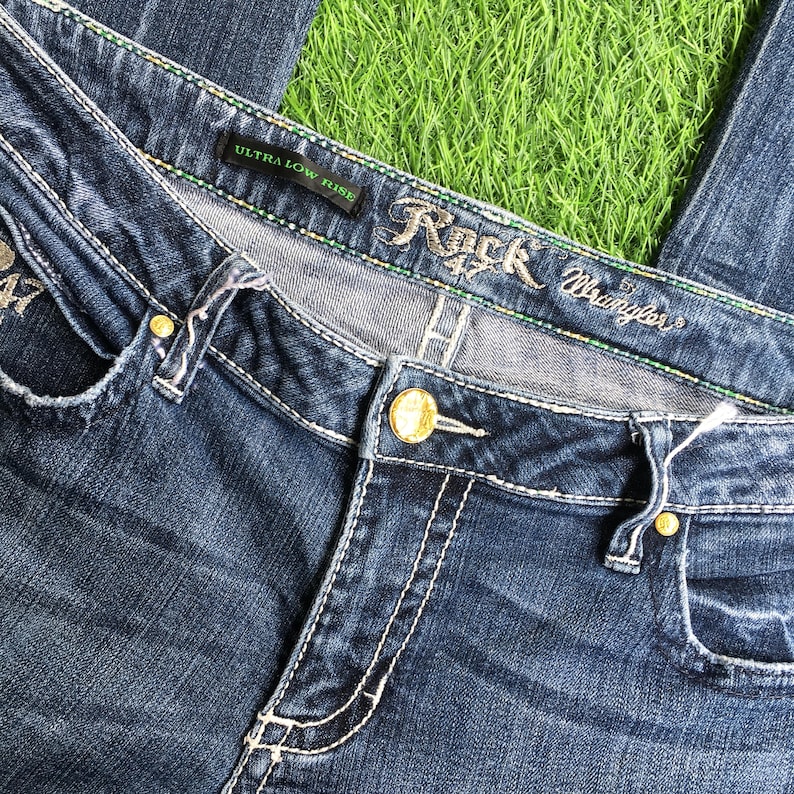 Op de afbeelding: Blauwe denim jeans met goudkleurige hardware en de tekst "Rock 47 by Wrangler" op de tailleband. De jeans zijn gemarkeerd met "Ultra Low Rise".