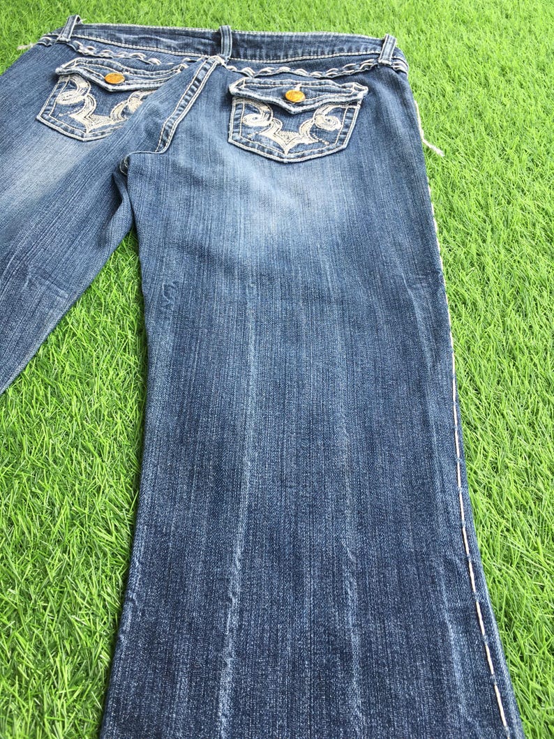 Op de afbeelding: Een paar blauwe denim jeans met witte stiksels en borduurwerk op de achterzakken. De jeans heeft een bootcut-stijl en is in goede staat.