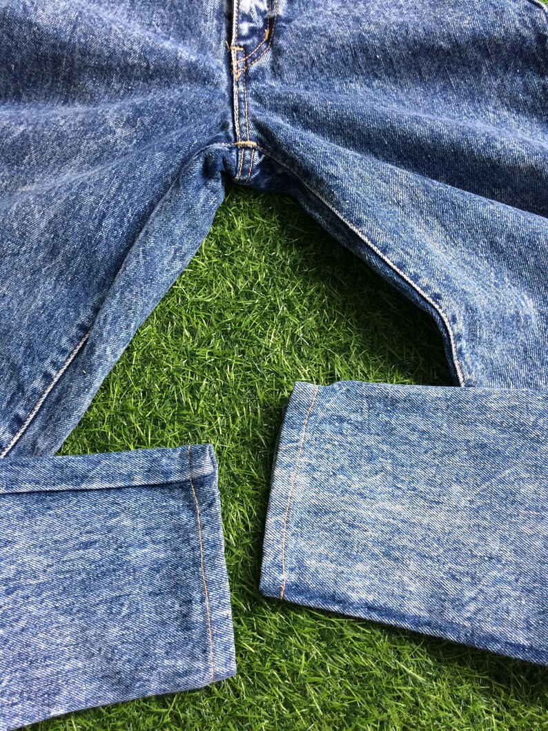 May include: A pair of blue denim jeans with a faded wash, laid out on a green grassy surface. The jeans have a straight leg and are cuffed at the bottom.