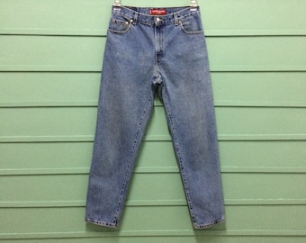 May include: Light blue denim jeans hanging against a green wooden wall. The jeans have a classic five-pocket design, a button and zipper closure, and a red label on the back. The jeans are a medium wash and appear to be in good condition.