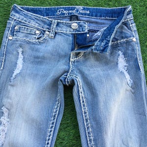 May include: Light blue denim jeans with distressed details and white stitching. The jeans feature a zipper fly, decorative rivets, and the brand name "Request Jeans" on the waistband. Size 5/27.
