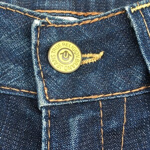 May include: Close-up of dark blue denim jeans with a gold button. The button is embossed with the words "TRUE RELIGION BRAND JEANS" around the edge and a horseshoe logo in the center. The jeans have orange stitching.