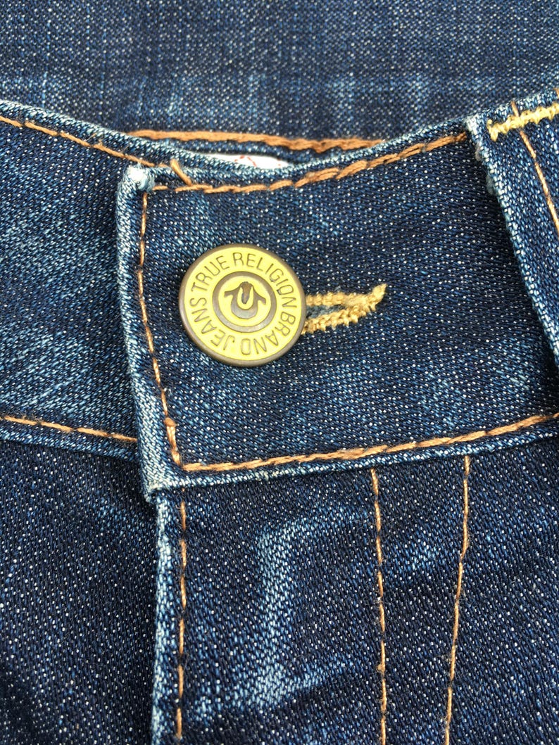 May include: Close-up of dark blue denim jeans with a gold button. The button is embossed with the words "TRUE RELIGION BRAND JEANS" around the edge and a horseshoe logo in the center. The jeans have orange stitching.