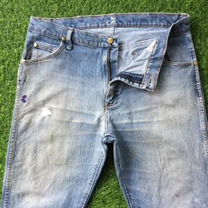 May include: A pair of light blue denim jeans with a faded look and a small hole on the left leg. The jeans have a zip fly and a button fastening. The waistband has a label that reads "VAI" in black text.