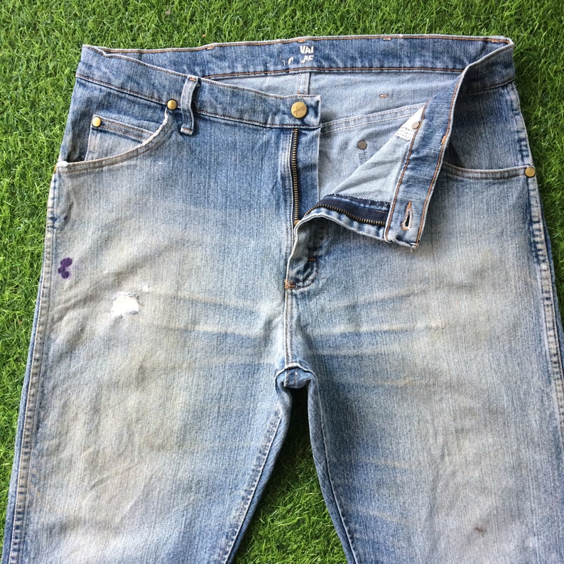 May include: A pair of light blue denim jeans with a faded look and a small hole on the left leg. The jeans have a zip fly and a button fastening. The waistband has a label that reads "VAI" in black text.