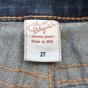 May include: Close-up of a denim fabric with a white label. The label reads "Religion - BRAND JEANS - Made in USA" and features the number "27". The stitching is visible in a contrasting brown color.