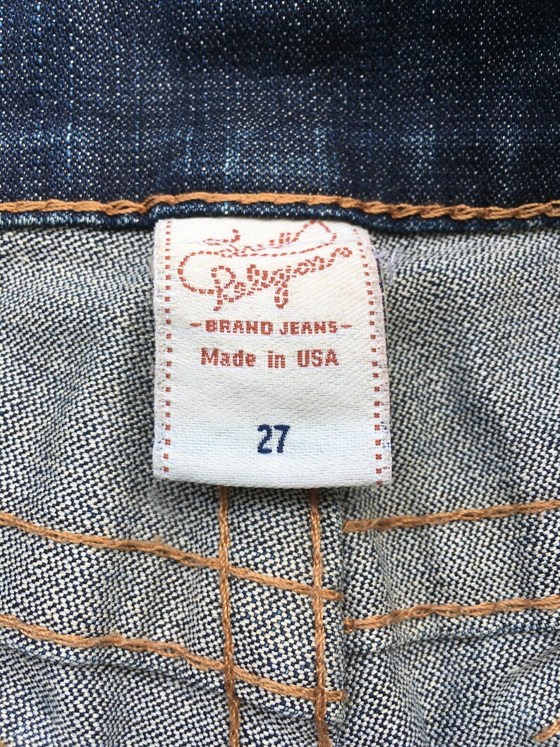 May include: Close-up of a denim fabric with a white label. The label reads "Religion - BRAND JEANS - Made in USA" and features the number "27". The stitching is visible in a contrasting brown color.
