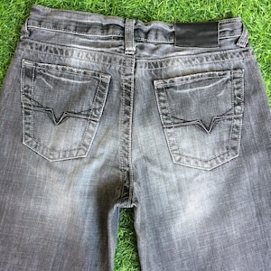 Può includere: Un paio di jeans in denim grigio scuro con due tasche posteriori e una toppa in pelle nera sul retro della cintura.