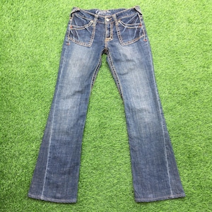 Puede incluir: Vaqueros de mezclilla azul con diseño de pernera acampanada. Los vaqueros tienen bolsillos delanteros, trabillas y costuras decorativas en naranja y rojo. Los vaqueros están colocados sobre una superficie verde.