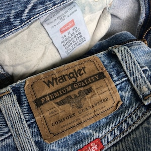 May include: A close-up of a pair of blue denim jeans with a brown leather Wrangler label that reads "Premium Quality" and "Comfort Guaranteed." The label also includes the text "Greensboro, North Carolina." The jeans have a red Wrangler label sewn on the bottom right corner.