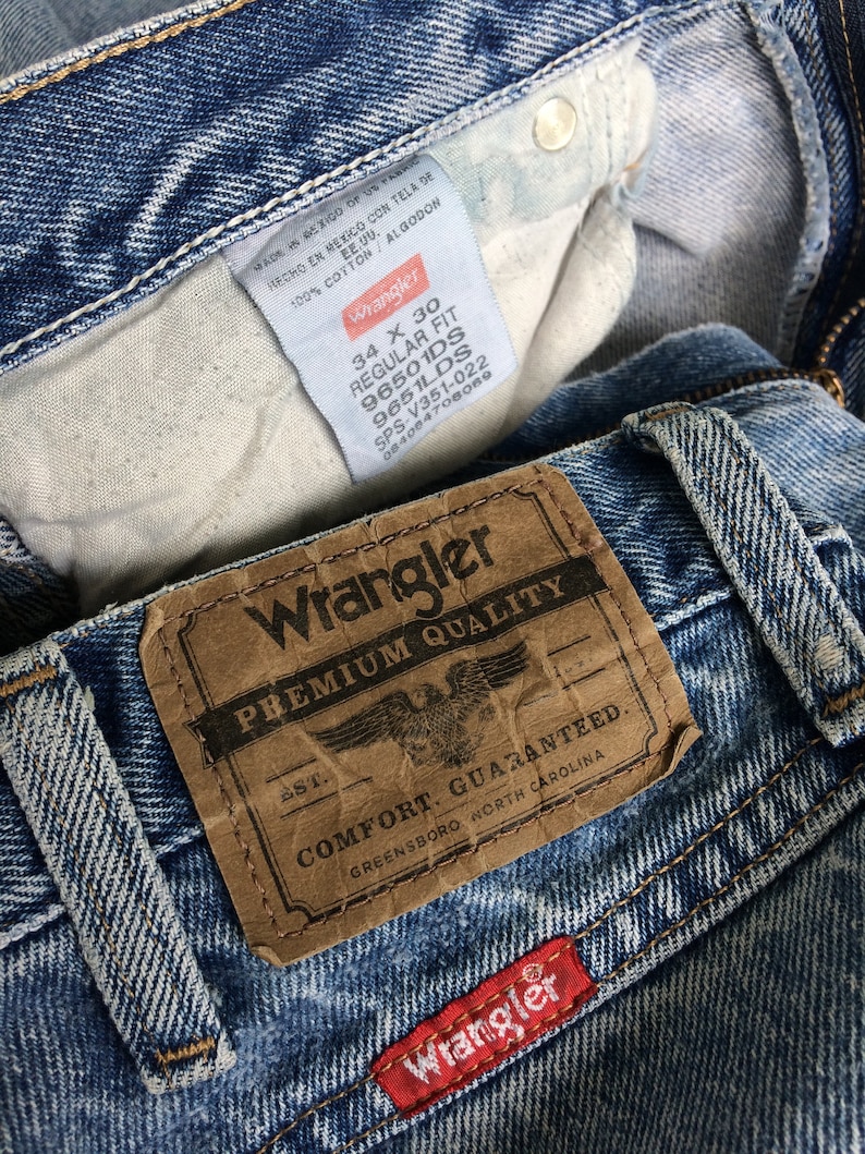 May include: A close-up of a pair of blue denim jeans with a brown leather Wrangler label that reads "Premium Quality" and "Comfort Guaranteed." The label also includes the text "Greensboro, North Carolina." The jeans have a red Wrangler label sewn on the bottom right corner.