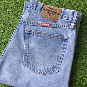 Pode incluir: Jeans de denim azul claro com um bolso traseiro e uma etiqueta Wrangler que diz "Premium Quality" e "Comfort Guaranteed".