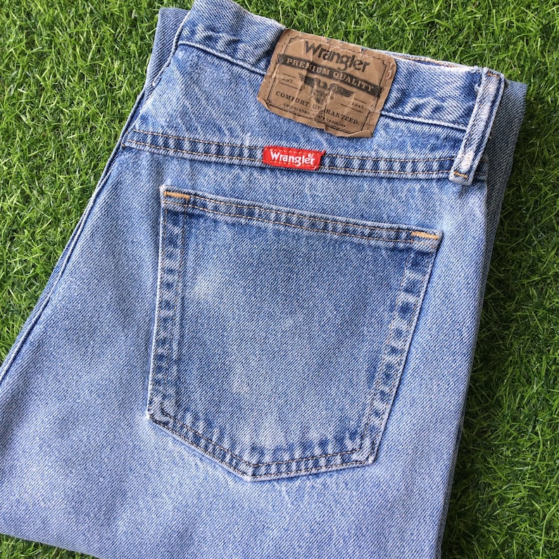 Pode incluir: Jeans de denim azul claro com um bolso traseiro e uma etiqueta Wrangler que diz "Premium Quality" e "Comfort Guaranteed".
