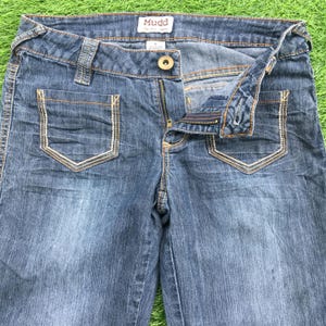 Puede incluir: Un par de pantalones vaqueros azules de mezclilla con cierre de cremallera y botón. Los vaqueros tienen un lavado claro y cuentan con un pequeño bolsillo a cada lado del frente. Los vaqueros tienen una etiqueta que dice "Mudd" en la cintura.