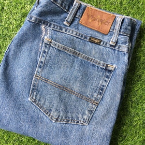 Pode incluir: Um par de calças de ganga azuis com um remendo de couro castanho que diz "Wrangler" no bolso de trás. As calças de ganga têm um bolso de trás com um design cosido.