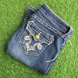 Op de afbeelding: Een paar blauwe denim jeans met een wit geborduurd zakdetail en een gouden knoop. De jeans is dubbelgevouwen en heeft een verweerde look.